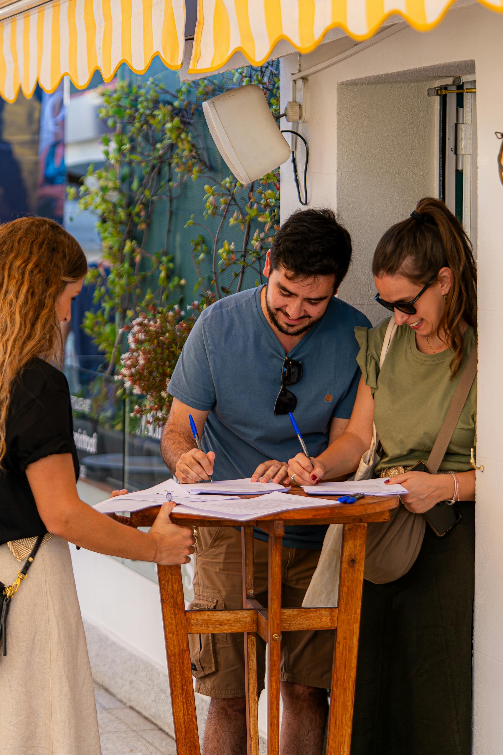 Three young adults signing documents at a table outside under a yellow awning.